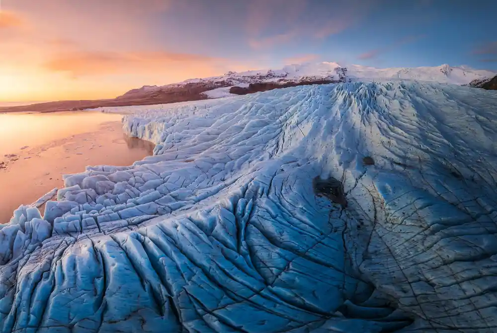 Glacier with blue ice formations at sunset, mountains beyond
